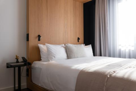 A double bed with wooden wall stands next to a window of a hotel room