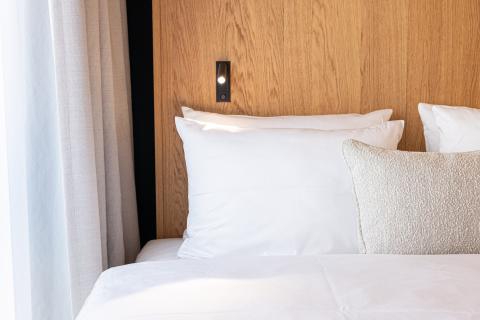 Aufgestellte Kissen in hellen Farben am Kopfende eines Hotelbettes mit Holzwand