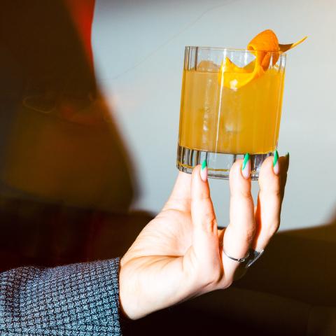 Eine Frau mit bunten Fingernägeln präsentiert einen gelben Cocktail